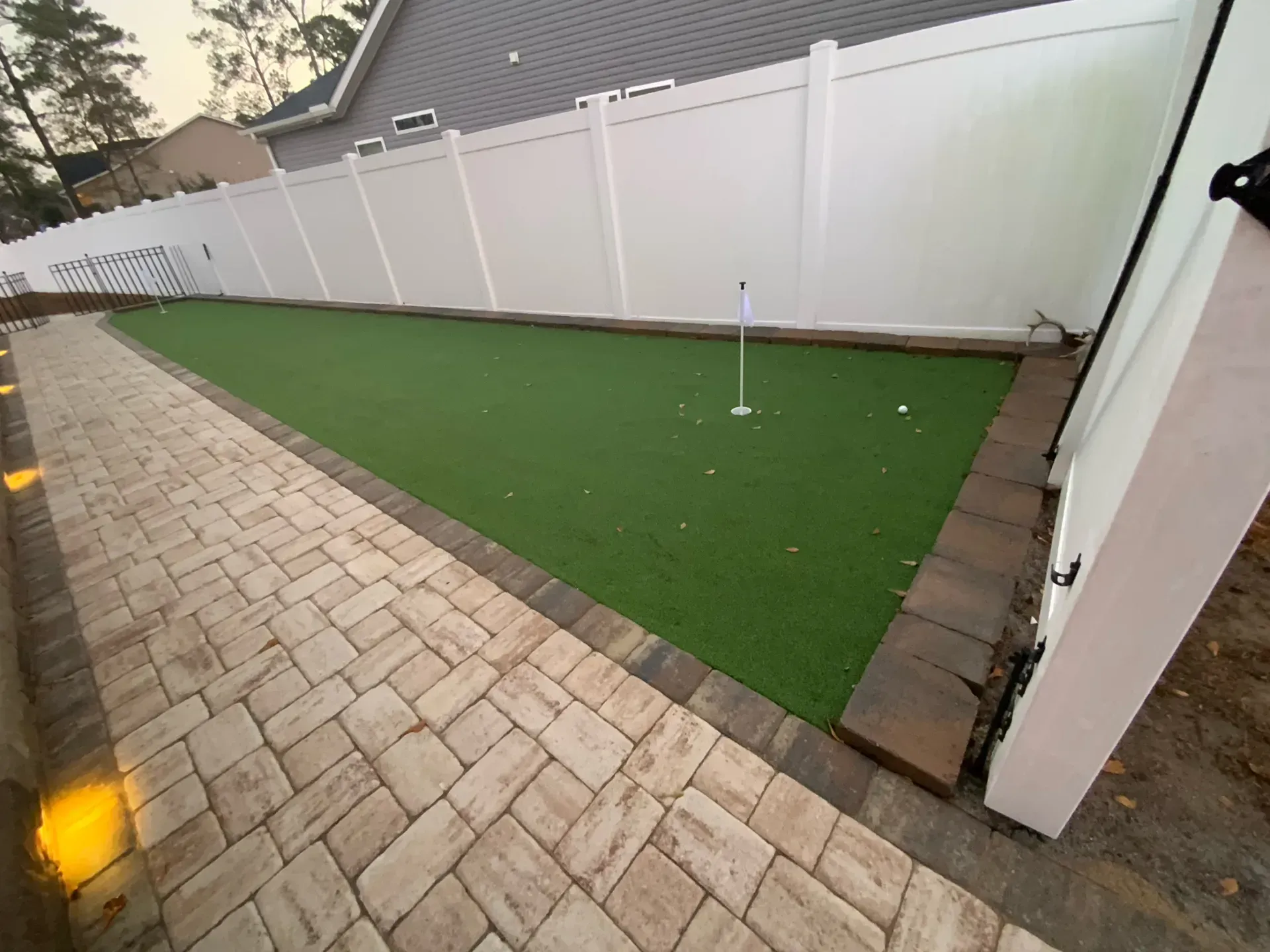 Backyard with white vinyl fence, artificial turf, brick pavers, and a golf putting cup.