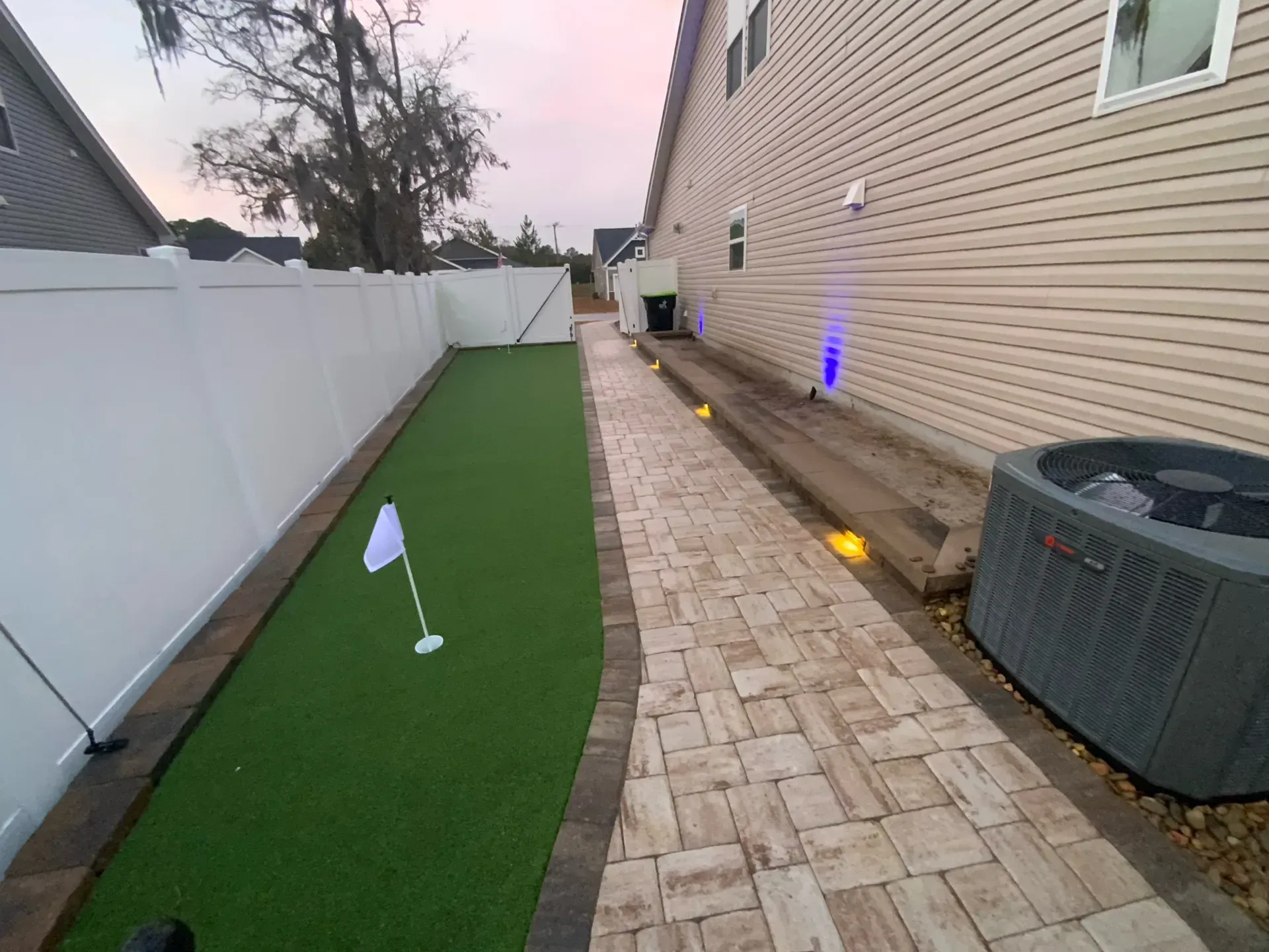 Narrow backyard with artificial turf, paver path, white fence, and house siding lit by small ground lights
