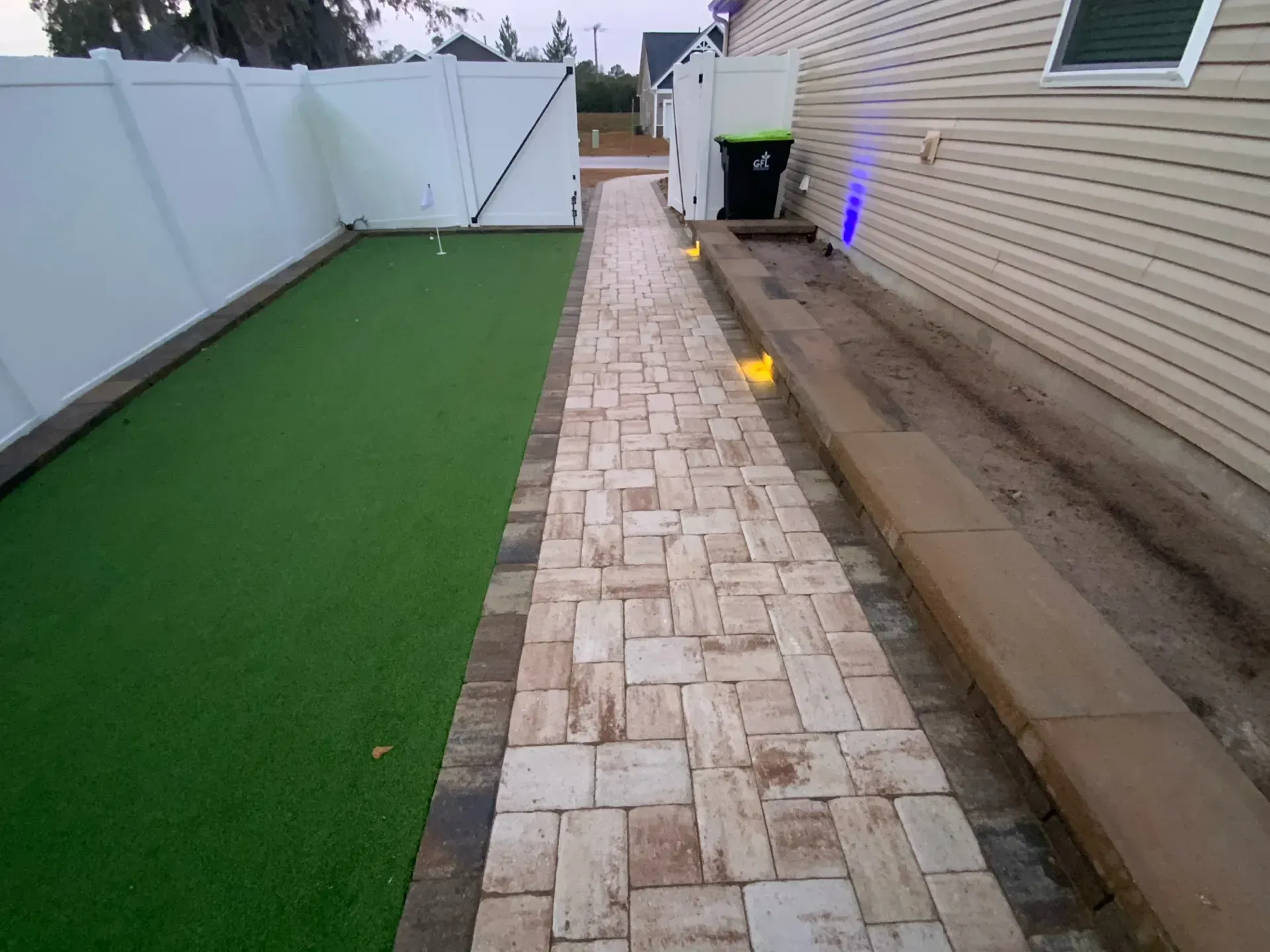 Narrow stone walkway beside a house, with white fence, green turf, and wet ground after rain.