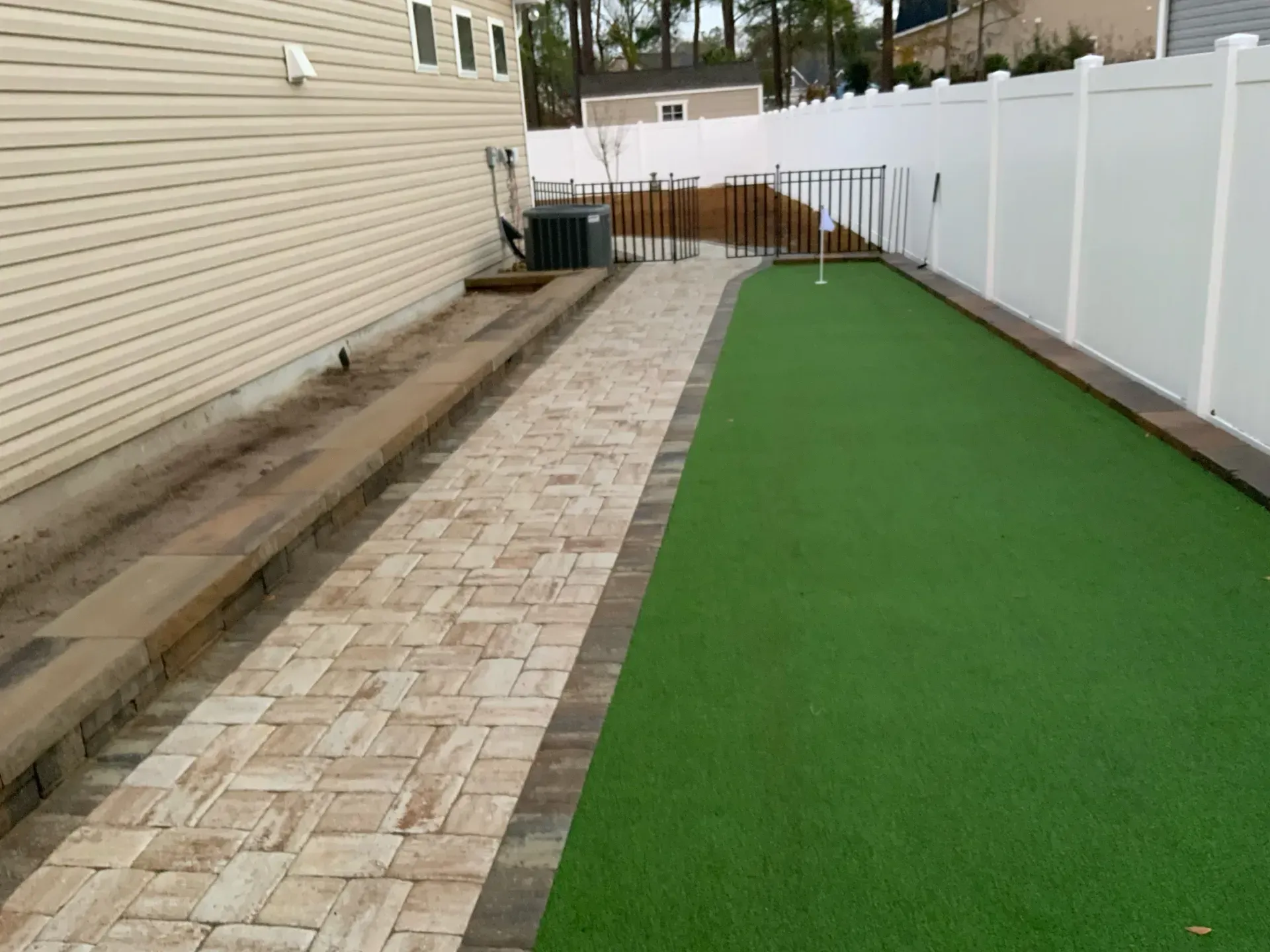 Narrow backyard path with brick pavers, green turf, beige house siding, and white vinyl fence.