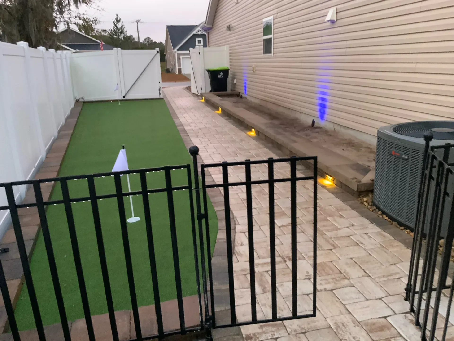Backyard with black gate, green turf, paved walkway, beige house, and white fence at dusk