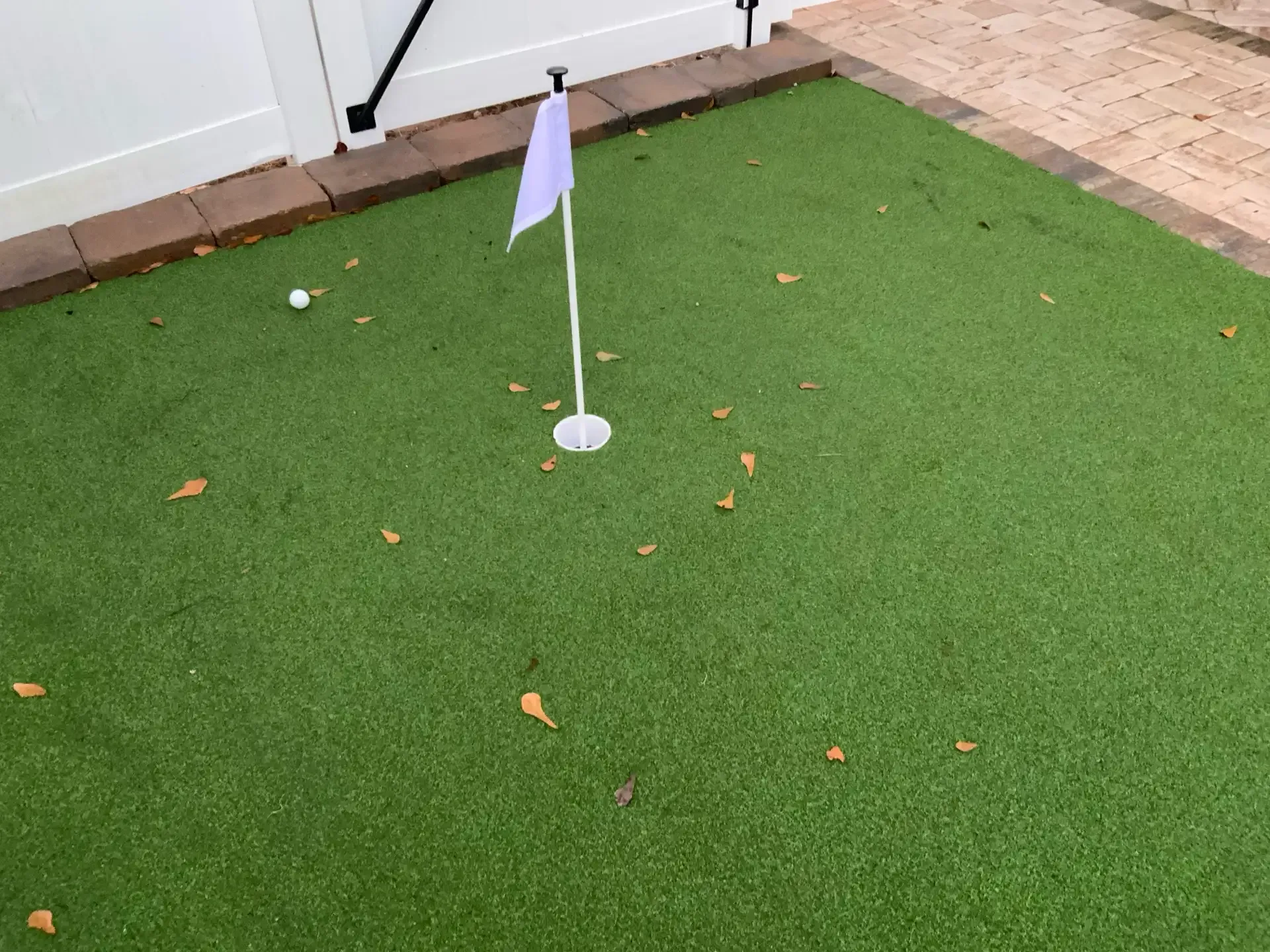 Small white flag on a putting green with scattered leaves near a patio wall