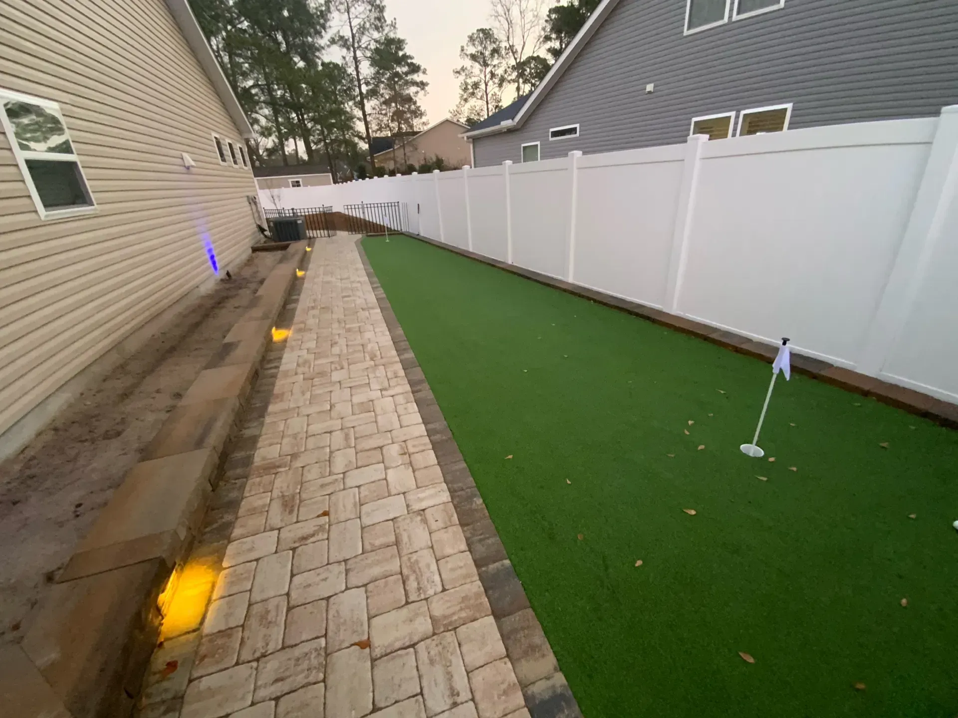 Narrow backyard walkway beside a white fence, with green turf and small ground lights along the path.