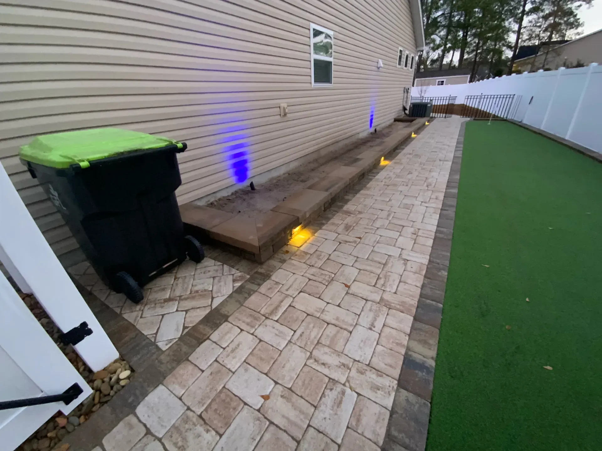 Narrow paver walkway beside a house, with blue lights, a wood deck, trash bins, and a white fence.