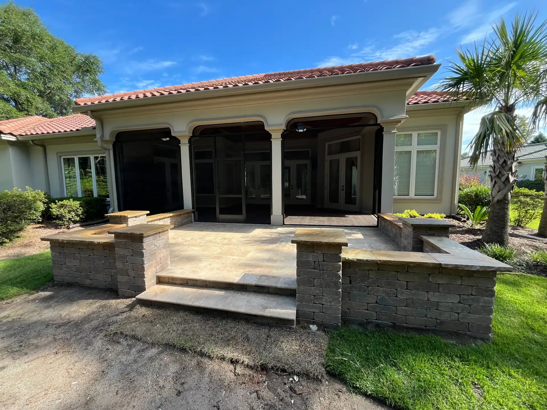 Front porch of a stucco house with stone steps, columns, and palm trees under a bright blue sky