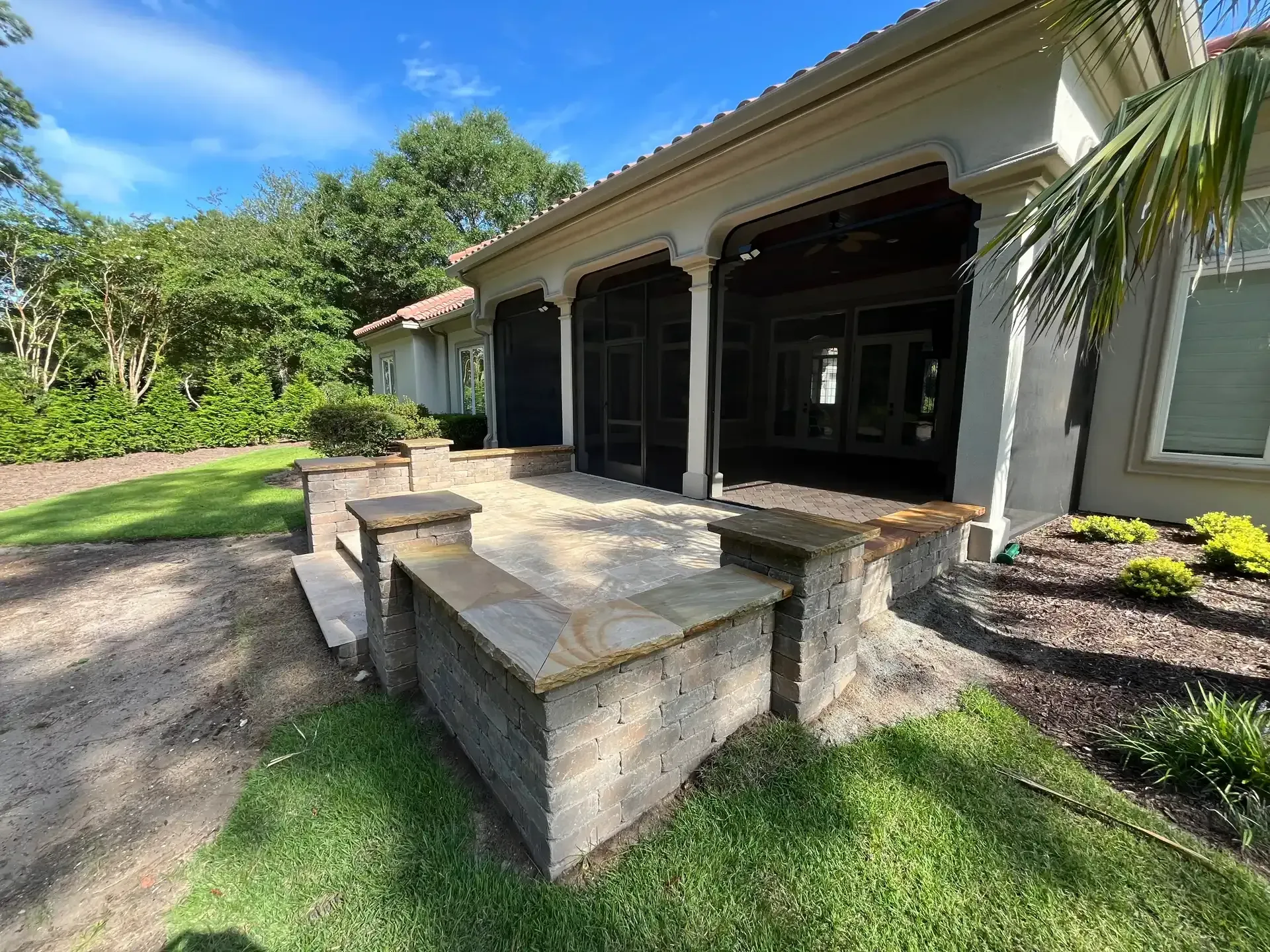 Stone patio with steps leading to an open patio beside a house, surrounded by green lawn and tropical plants