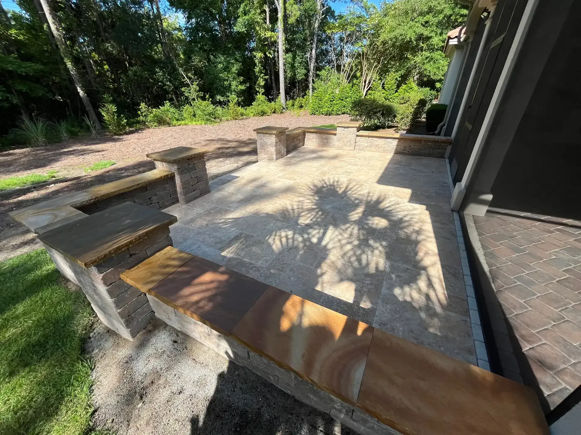Wooden patio benches around a sunlit concrete patio beside a dark house, with trees in the background