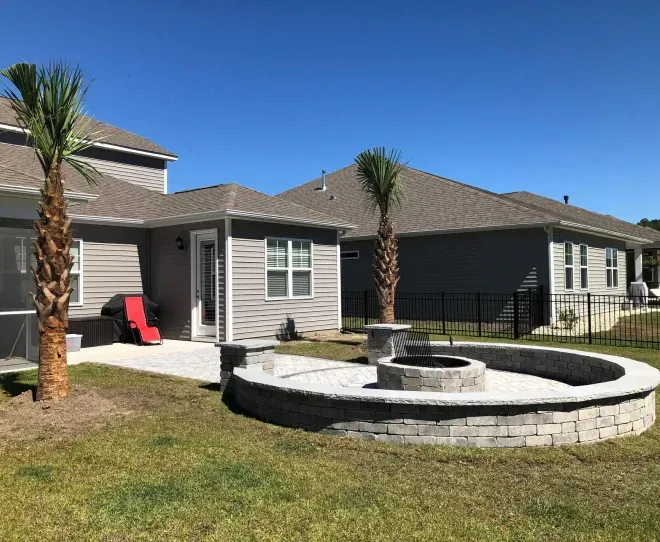 Suburban backyard with stone fire pit, two palm trees, and a patio beside a gray house under a clear blue sky