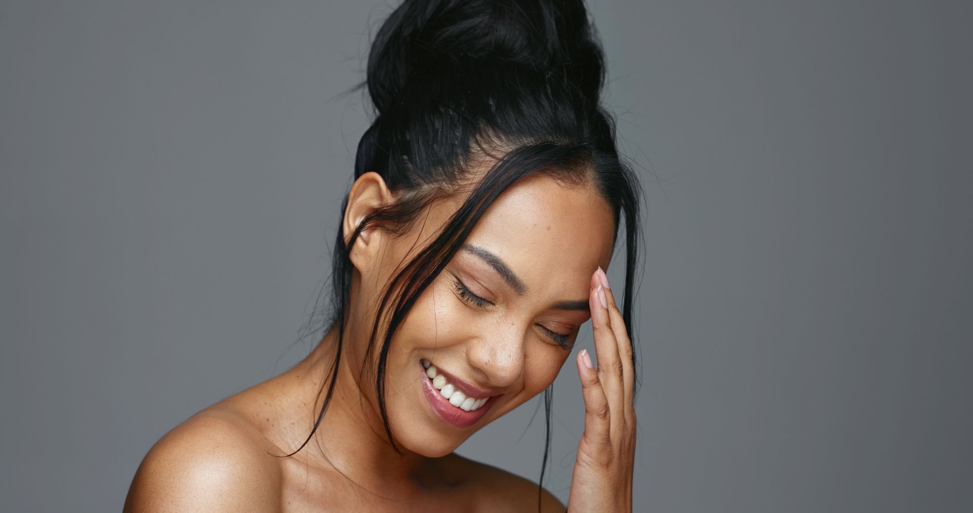 Smiling woman with dark hair touching her face against a gray background