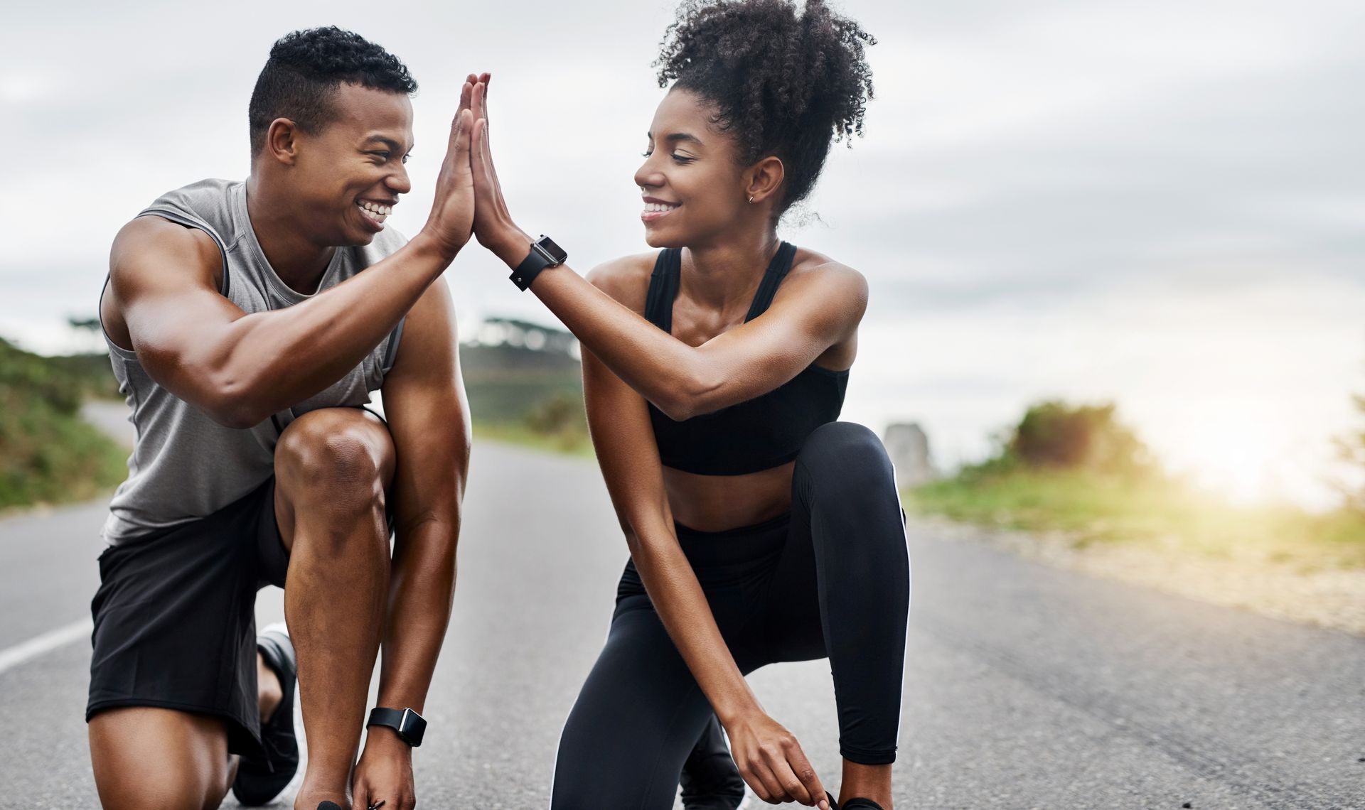 Two runners crouch on a road and high-five, smiling before a workout.