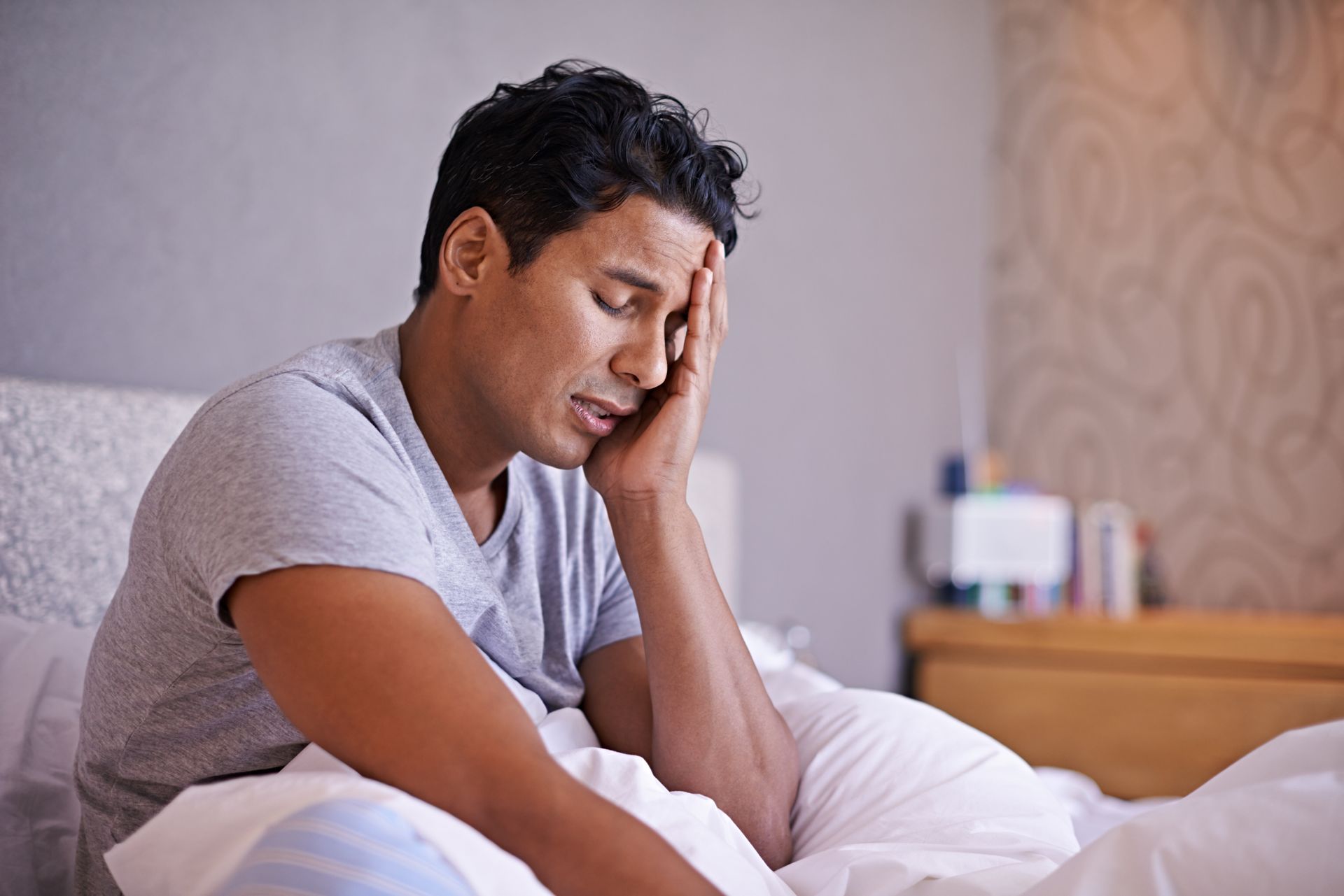 Man sitting on a bed, holding his forehead with a pained expression in a bedroom