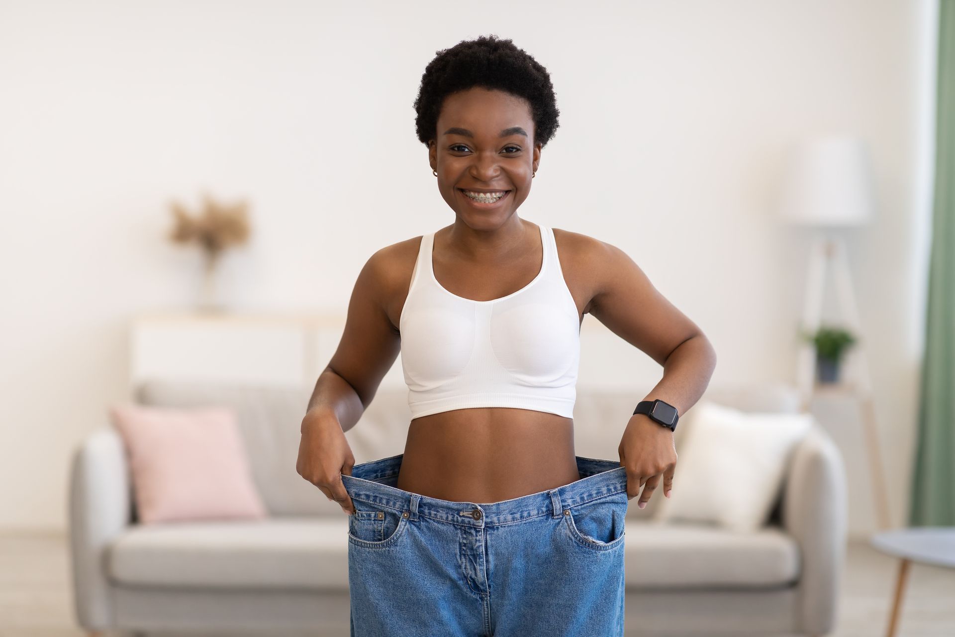 Person in white sports bra and jeans, standing in a bright living room and pulling up oversized pants.
