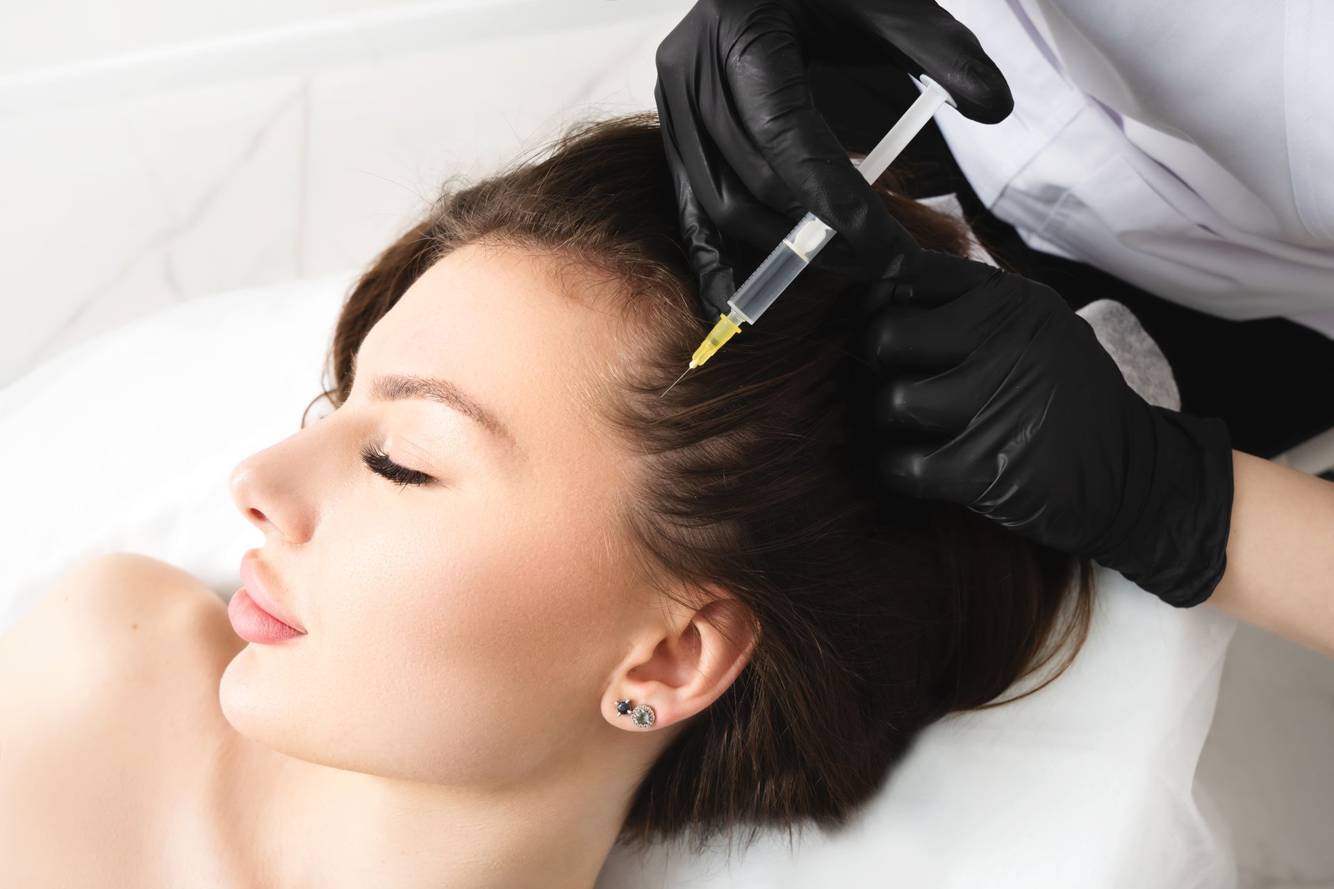 Woman receiving a scalp injection for hair loss, by a person wearing black gloves, in a medical setting.
