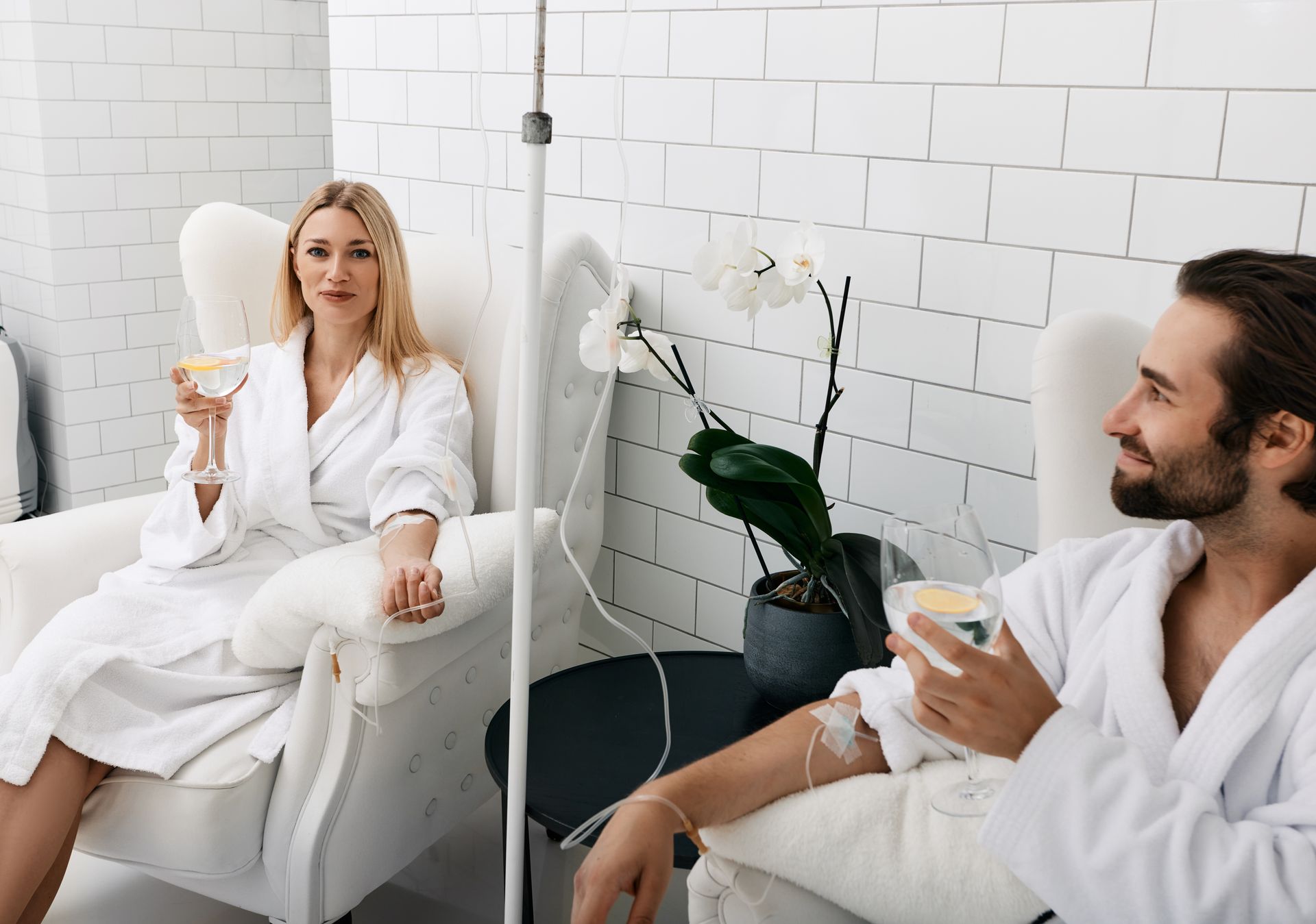 Two people in white robes sit in a bright spa lounge, each holding a glass of wine.
