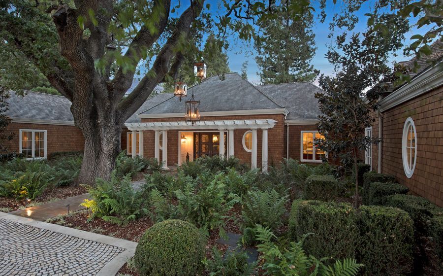 Brick house entrance with lit windows, surrounded by lush trees and garden at dusk