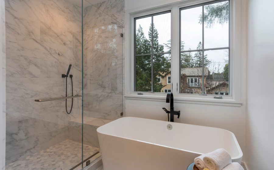 Modern bathroom with freestanding tub, glass shower, and large window overlooking trees and houses