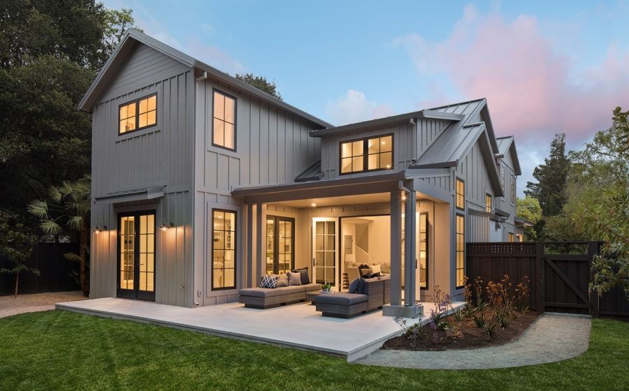 Modern two-story gray house with warm lights, large windows, and a backyard patio at dusk