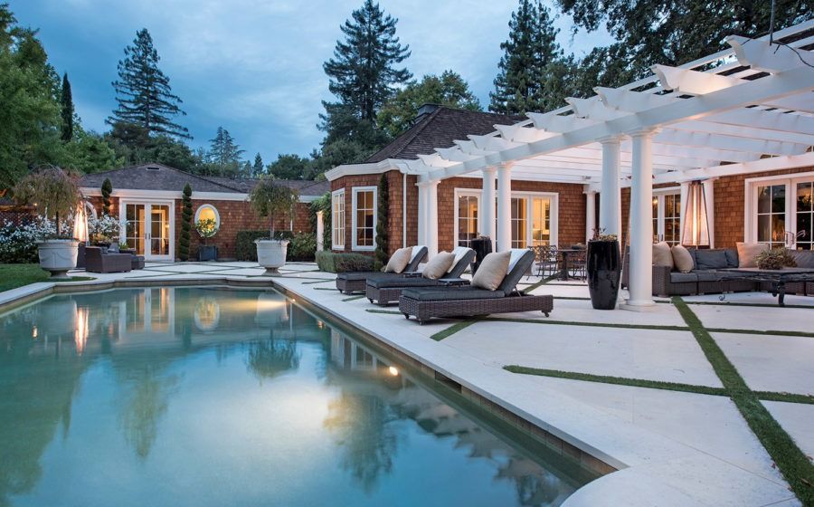 Luxury backyard pool at dusk with lounge chairs, pergola, and modern house glowing in warm light
