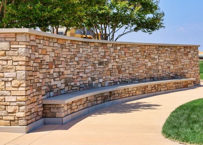 Curved stone bench against a tan stone wall beside a sidewalk and grass in a sunny outdoor park area