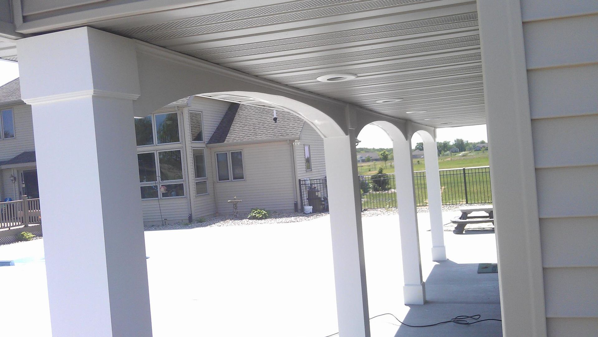 Covered patio with white columns and patterned ceiling beside a house in bright sunlight