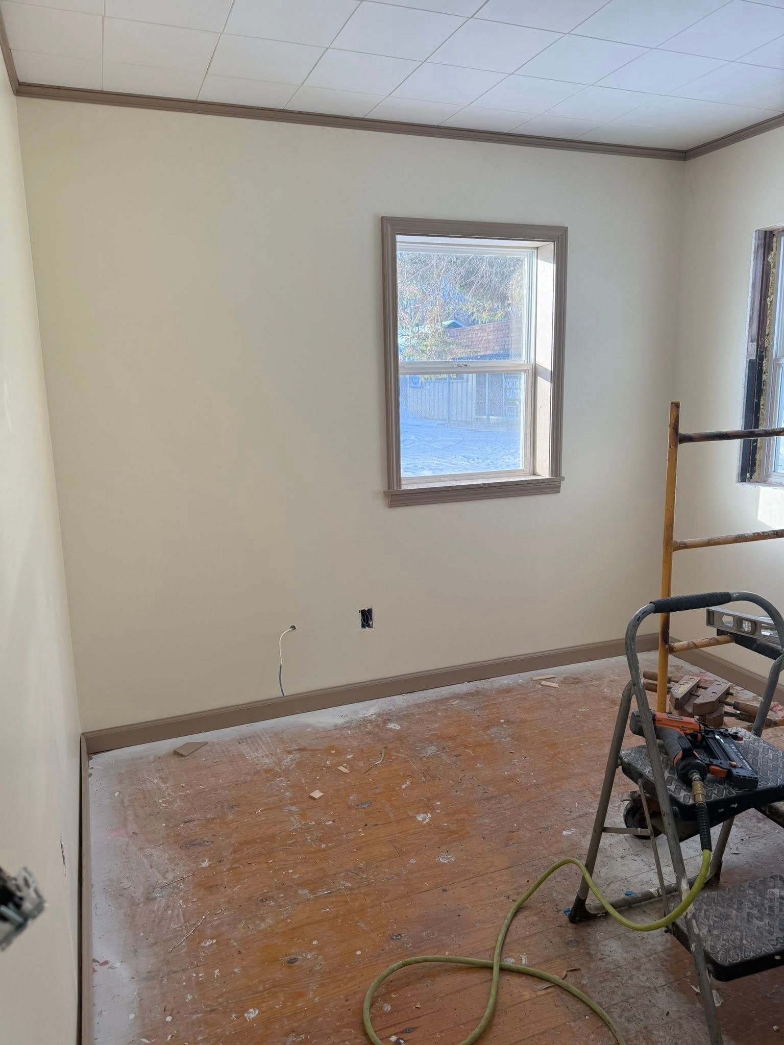 Small empty room with beige walls, a window, exposed subfloor, and a power tool on the right.