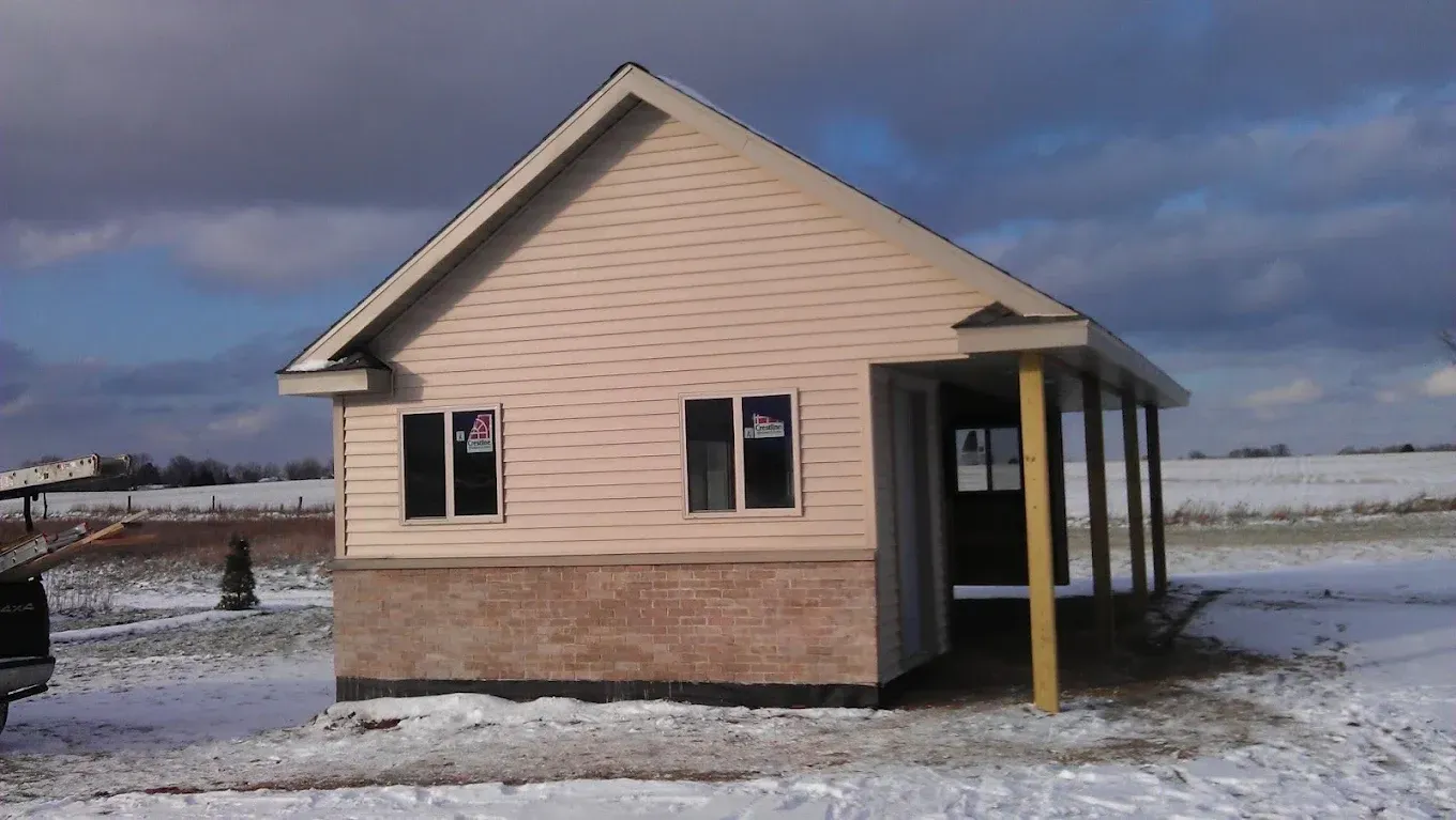 Small beige house with a porch in a snowy field under a cloudy sky