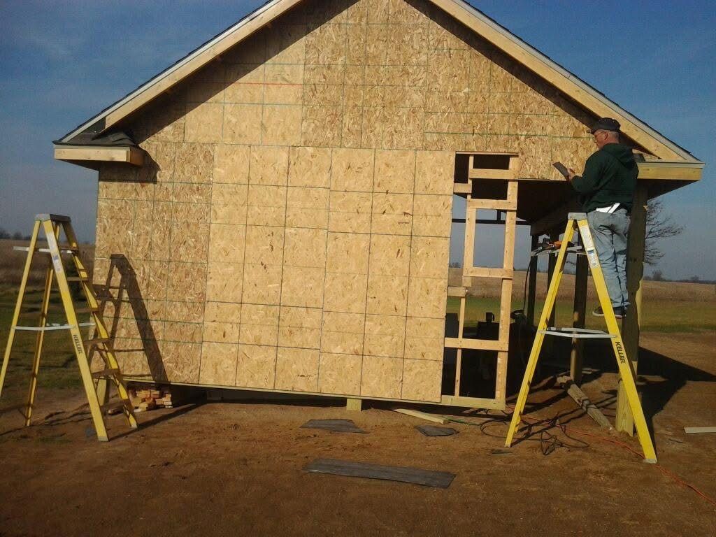Workers on ladders install stone veneer siding on a house exterior under construction.