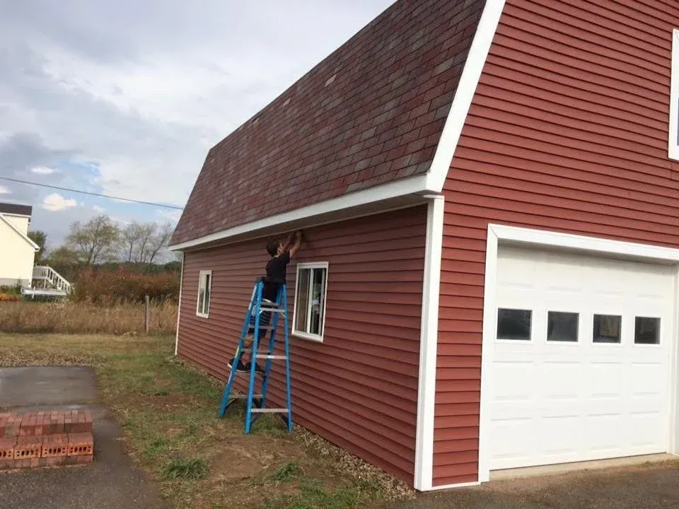 Person on blue ladder working on window of red house beside white garage door
