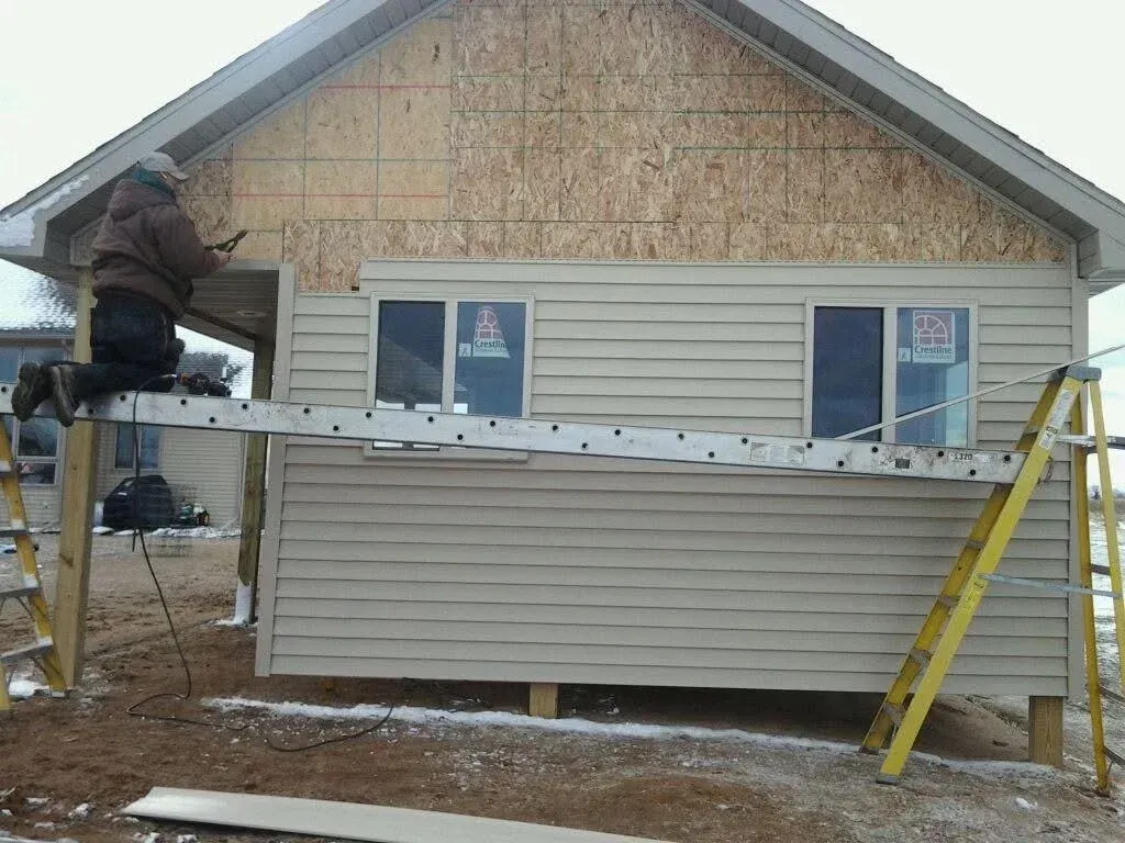 Worker on scaffolding installing siding on a house with plywood and beige exterior walls.