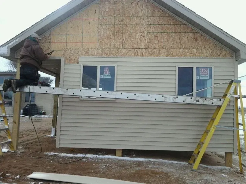 Worker on scaffolding installing siding on a house with plywood and beige exterior walls.