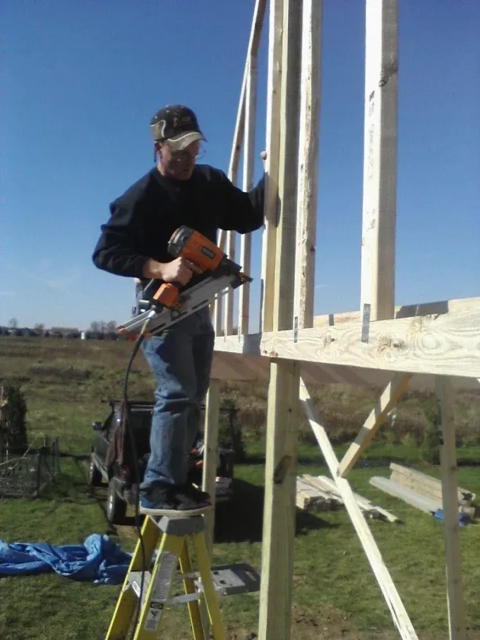 Person using a drill while standing on a step stool, building a wooden frame outdoors.