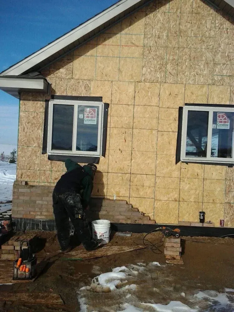 Person digging beside a tan house in snowy yard, near windows and a white bucket.