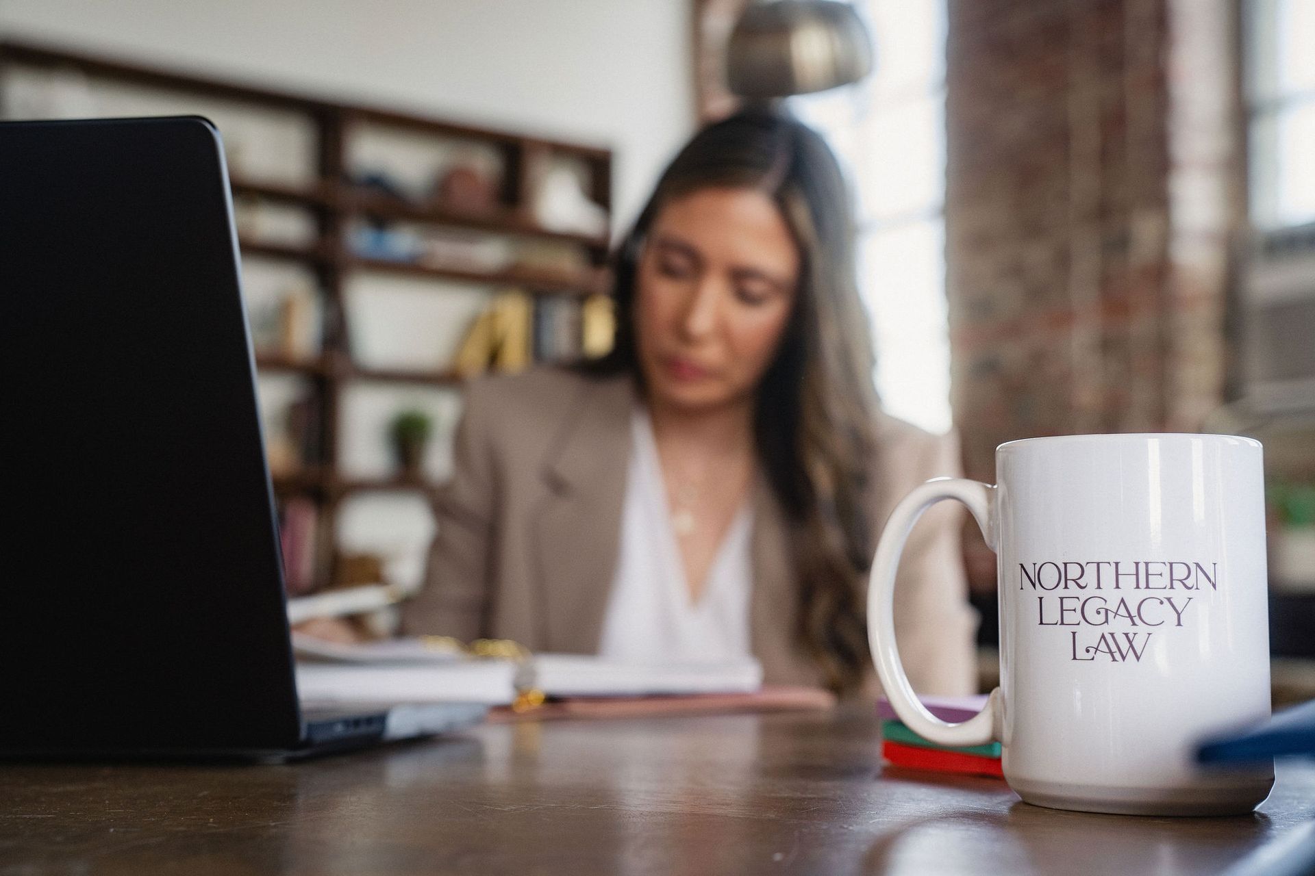 Woman working at a desk with a laptop, papers, and a white coffee mug in a blurred office setting