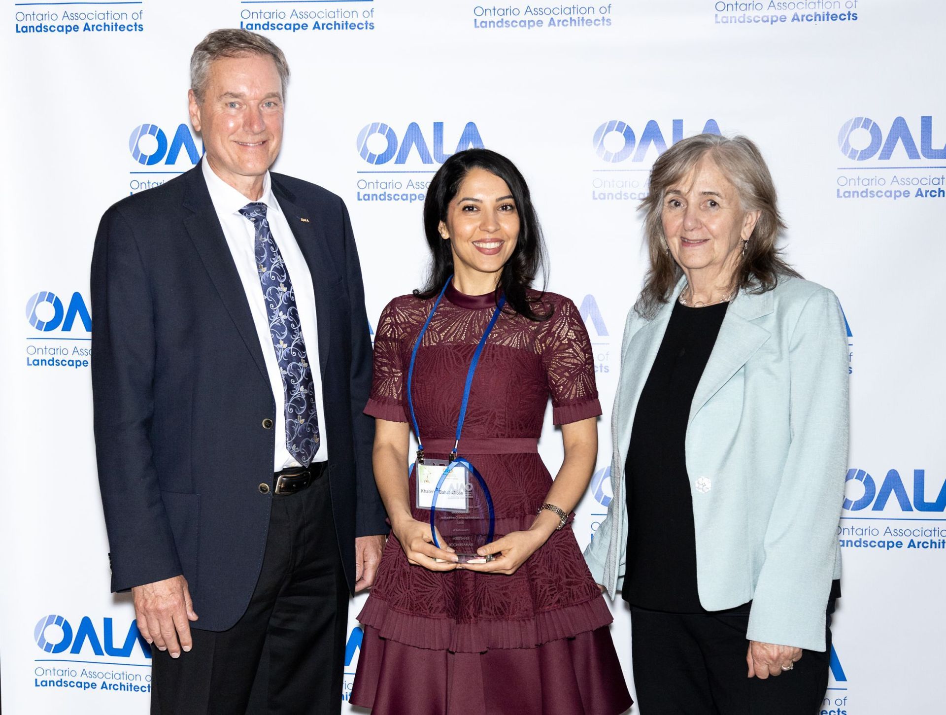 Three people posing at an OA event backdrop, with a woman centered holding an award plaque.
