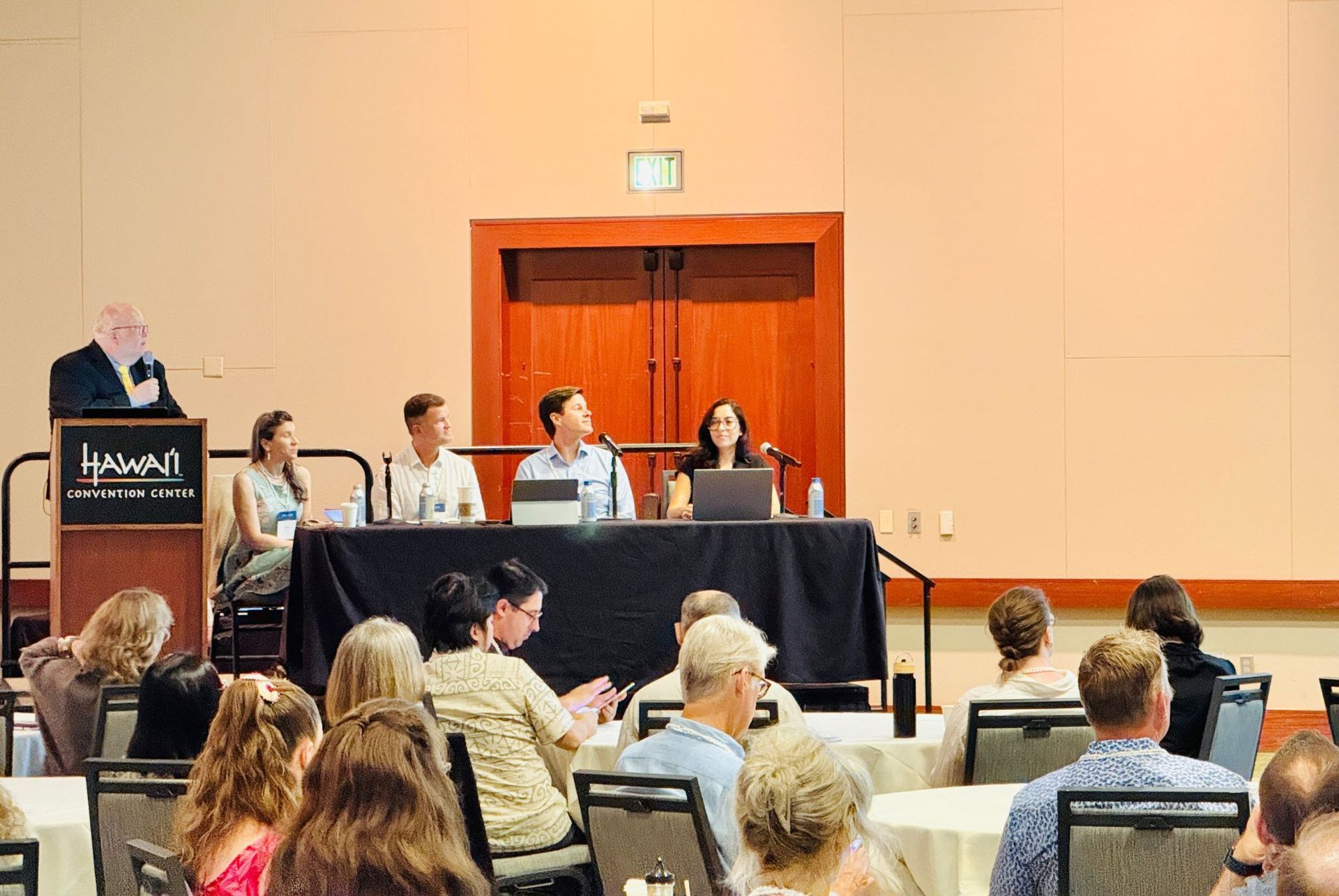 Panel discussion in a conference room with speakers seated at a long table and audience members watching.