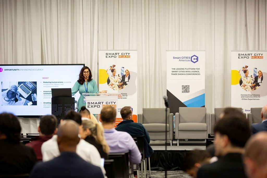 Conference speaker presenting on stage beside seated audience, with presentation banners and chairs in the background