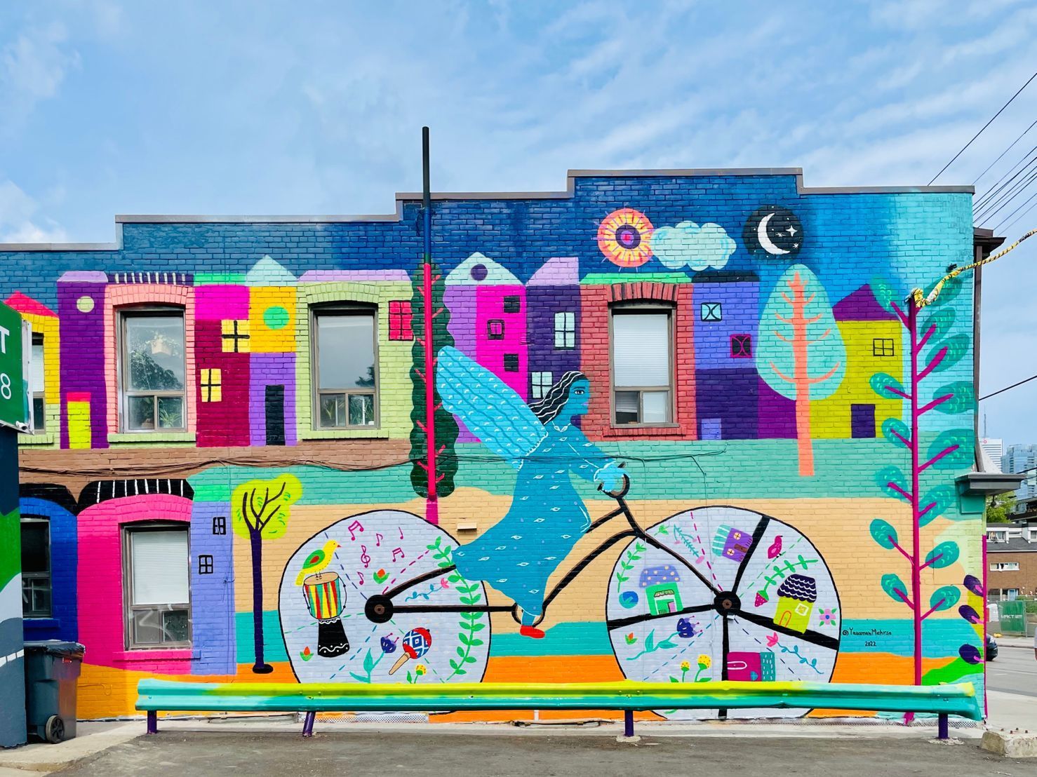 Colorful mural on a building with bright abstract shapes, bicycles, birds, and a blue sky backdrop