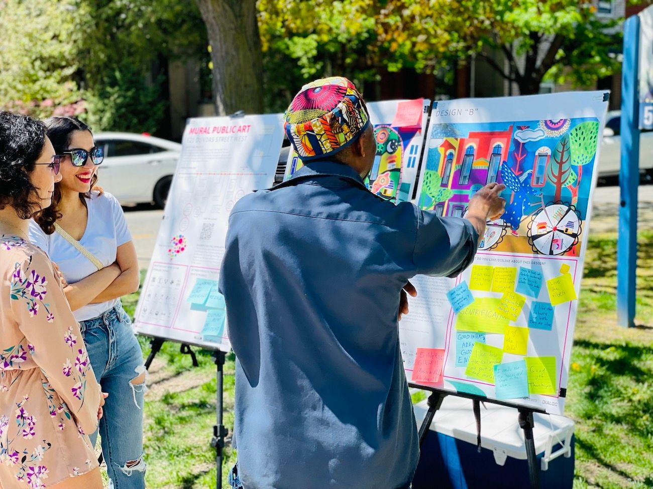 Artist painting at an outdoor easel while two people watch colorful canvases nearby.