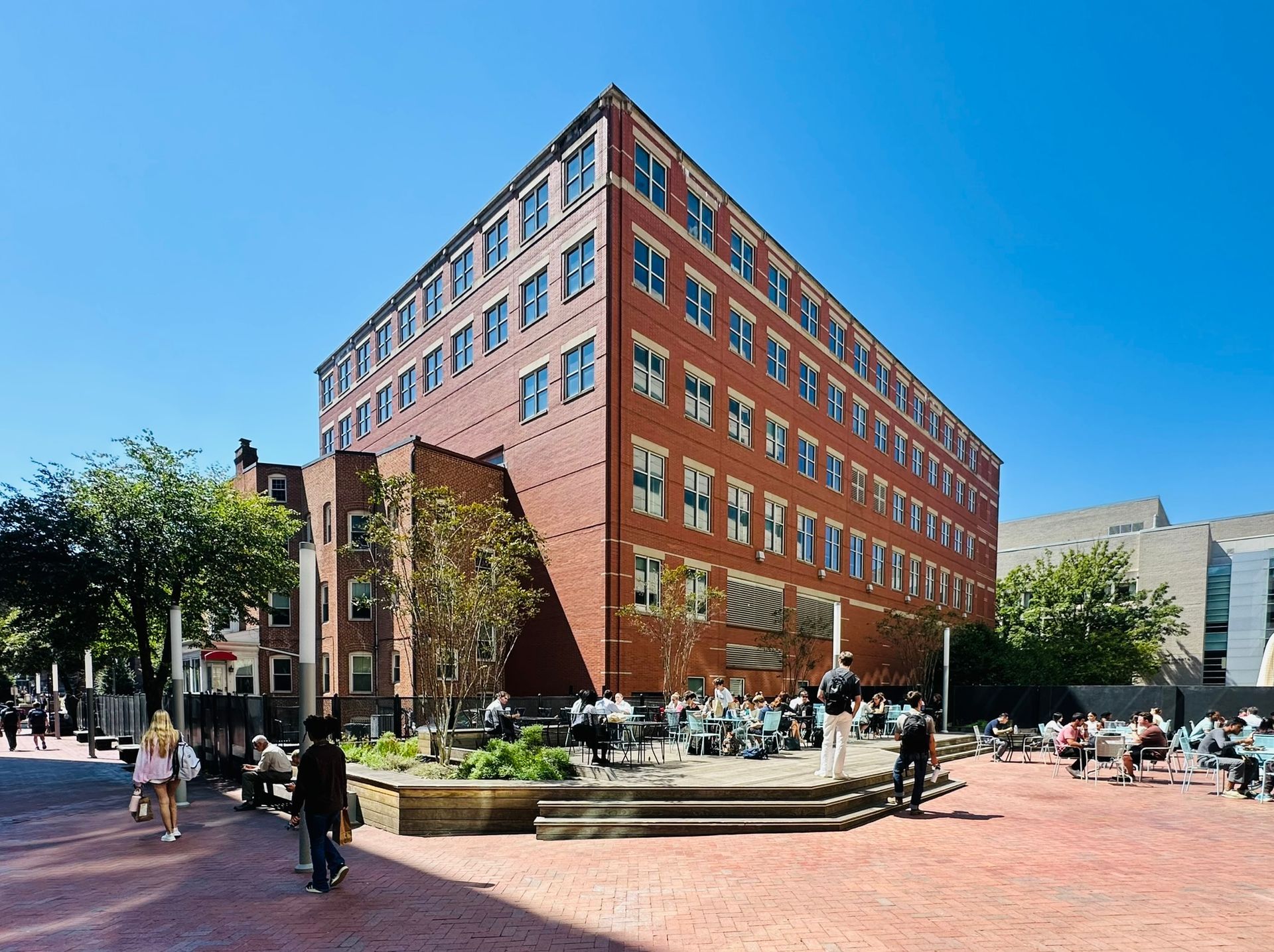 Brick office building on a sunny city corner with pedestrians, trees, and a red-brick plaza