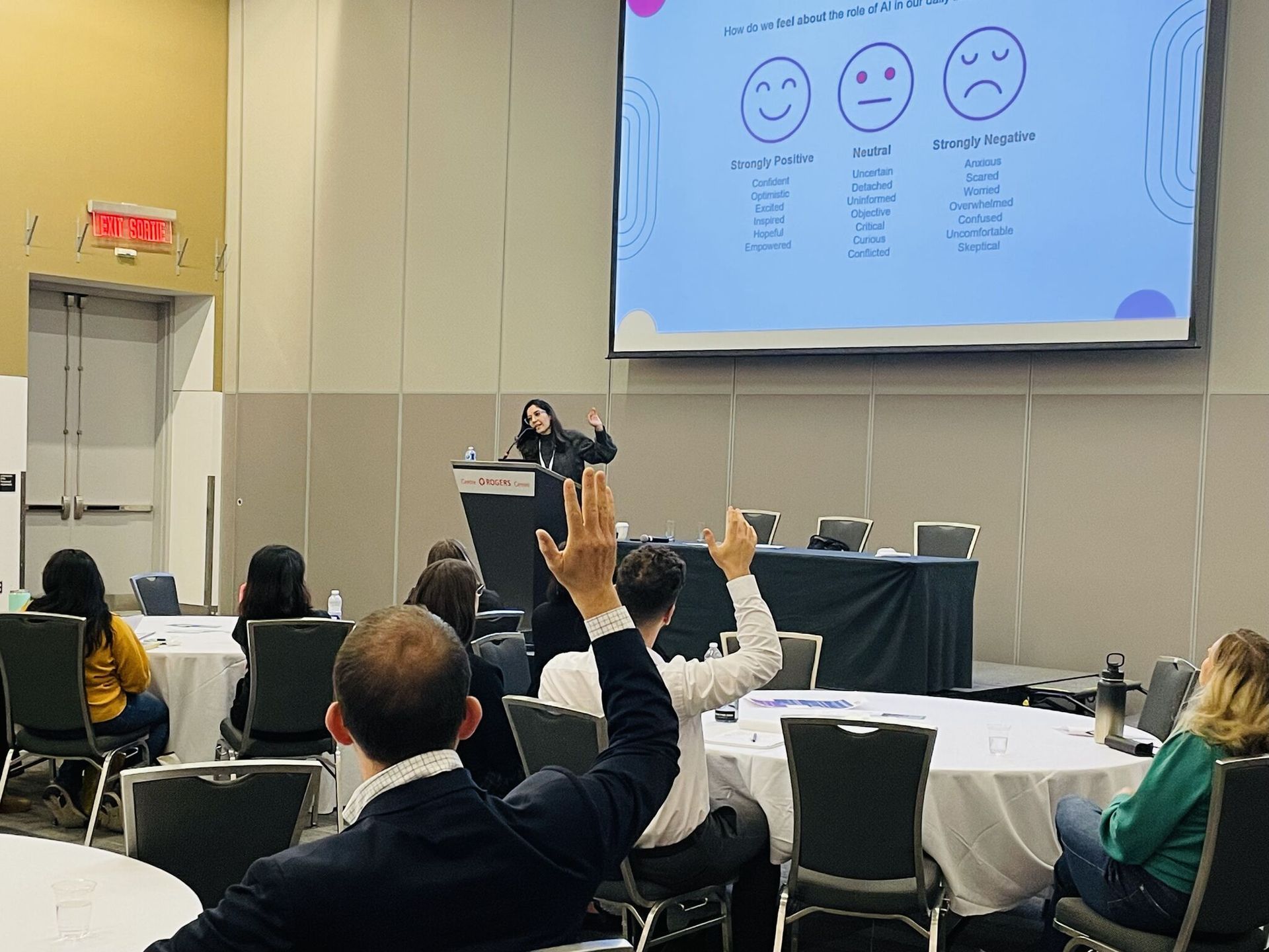 Audience raising hands during a presentation in a conference room with a slide showing smiley-face ratings.