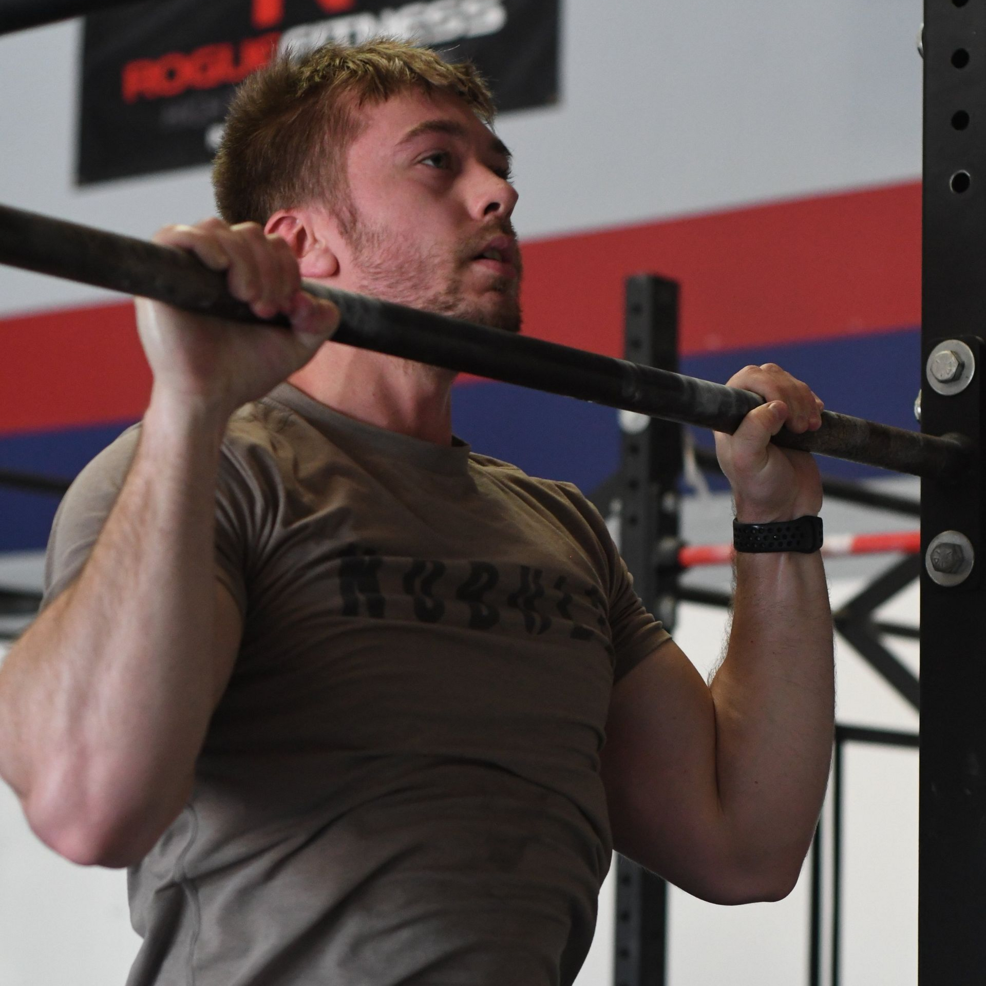 Man doing pull-ups on a bar in a gym, wearing a brown shirt and focused expression at Perry Hall Fitness & Performance in White Marsh, MD