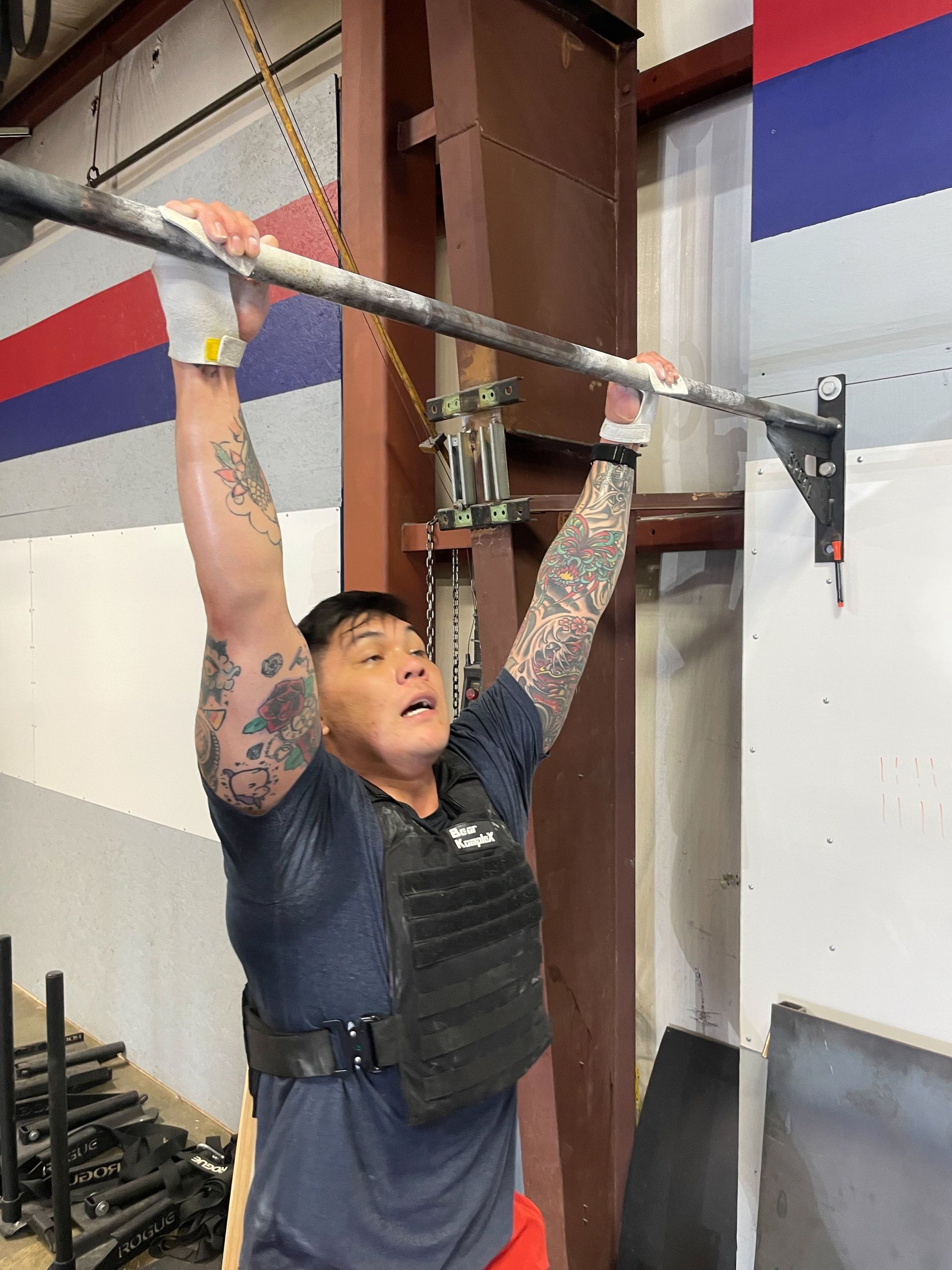 Person doing a pull-up on a bar in a gym, wearing a weighted vest and tattoos visible at Perry Hall Fitness & Performance in White Marsh, MD