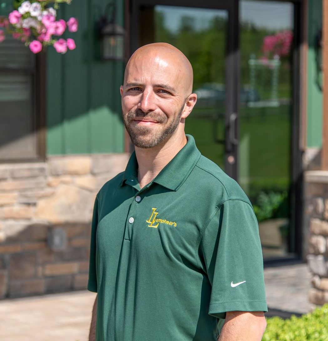 Man in green polo shirt smiles in front of a building with flowers and a glass door.