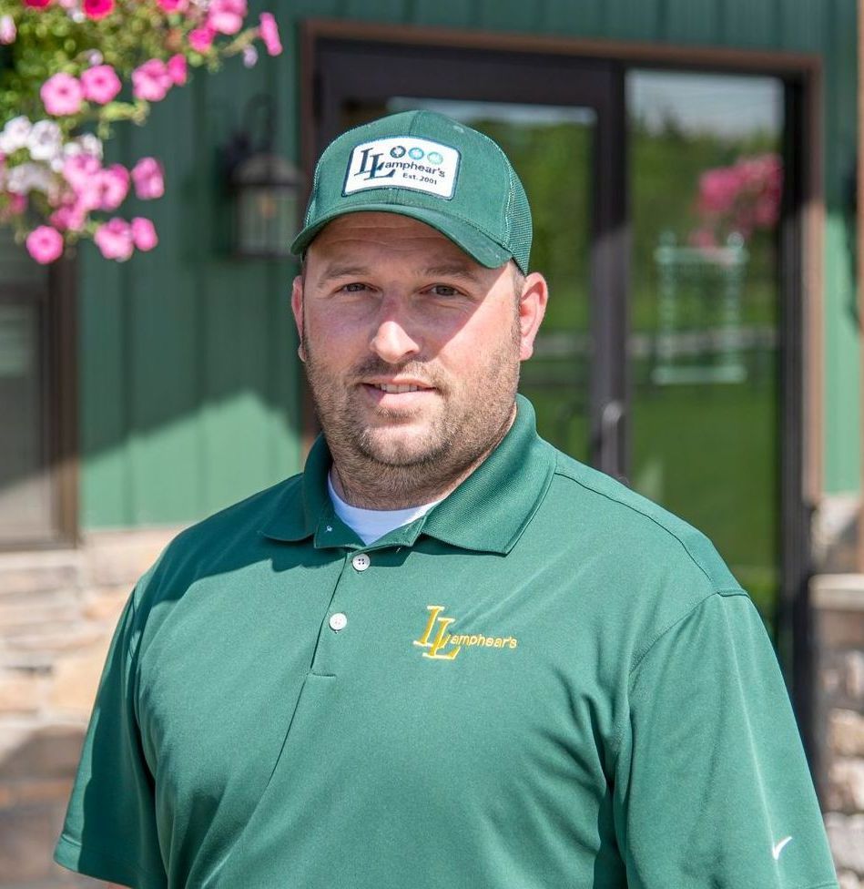 Man in green polo and cap, smiling, in front of a green building with flowers.