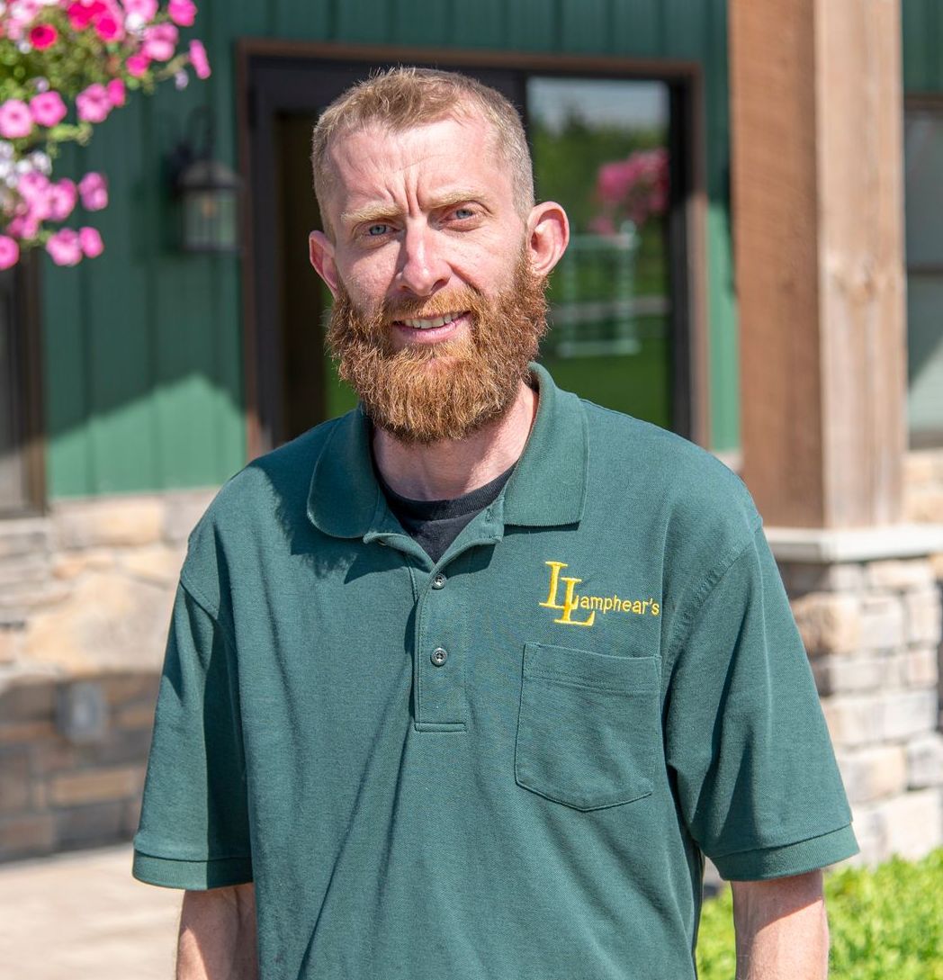 Man with a red beard in a green polo shirt, standing in front of a green building with pink flowers.