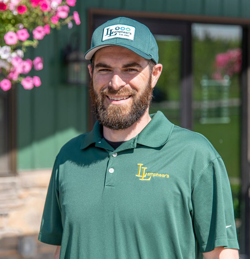 Man in green shirt and cap, smiling outdoors, flowers and green building in background.