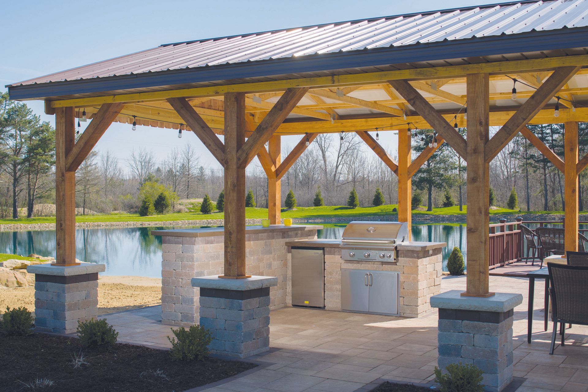 Outdoor kitchen with grill, seating, and pond view, under a wooden-framed shelter with metal roof.