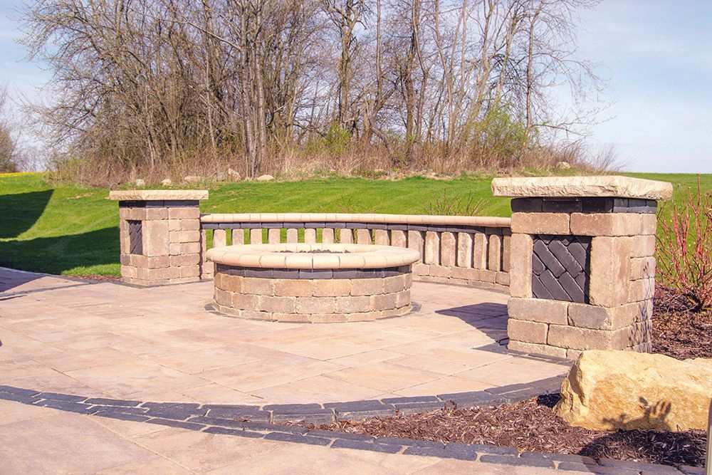 Brick-paved outdoor seating area with a low wall, pillars, and a circular fire pit on a lawn.