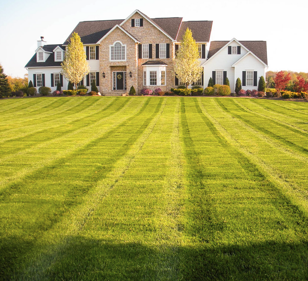 Two-story house with manicured green lawn, stripes of light and dark green, surrounded by trees.