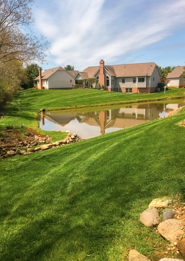 Lush green lawn slopes to a pond, reflecting houses with brick chimneys under a partly cloudy sky.